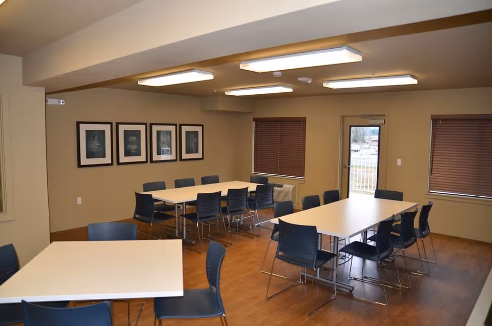 A well-lit room with three rectangular white tables surrounded by black chairs on a wooden floor. The walls are beige with four framed pictures hanging on one side. There are two windows with brown blinds and a glass door leading outside.