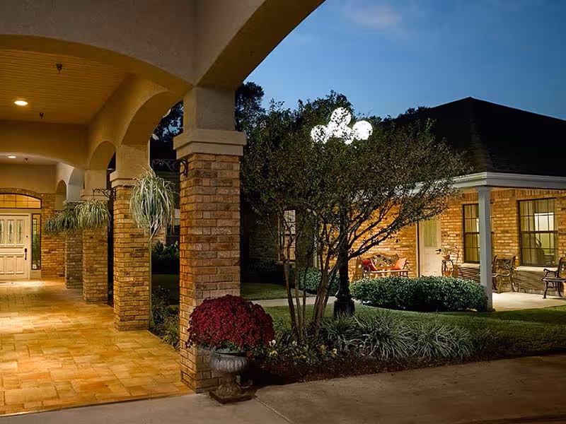 Evening view of the exterior of a senior living facility with brick columns and arches along a covered walkway, outdoor plants, a tree with a street lamp, and a seating area on a porch.