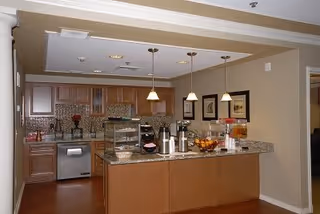 Interior view of a kitchen area with wooden cabinets, granite countertops, and stainless steel appliances. The kitchen island has coffee dispensers, a bowl of fruit, and a display case with baked goods. Three pendant lights hang above the island, and framed artwork is on the wall in the background.