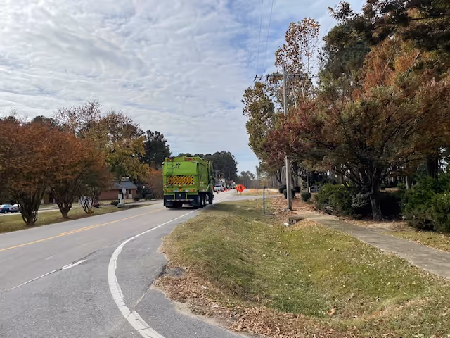 A green garbage truck driving down a tree-lined suburban road with fall foliage and a sidewalk.