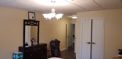 Interior view of a room in Woodland Ridge Assisted Living showing a dresser with a mirror, a chandelier light fixture, a closed double door, and a hallway leading to another room. There are some items on the dresser and a black trash bag on the floor near the hallway.