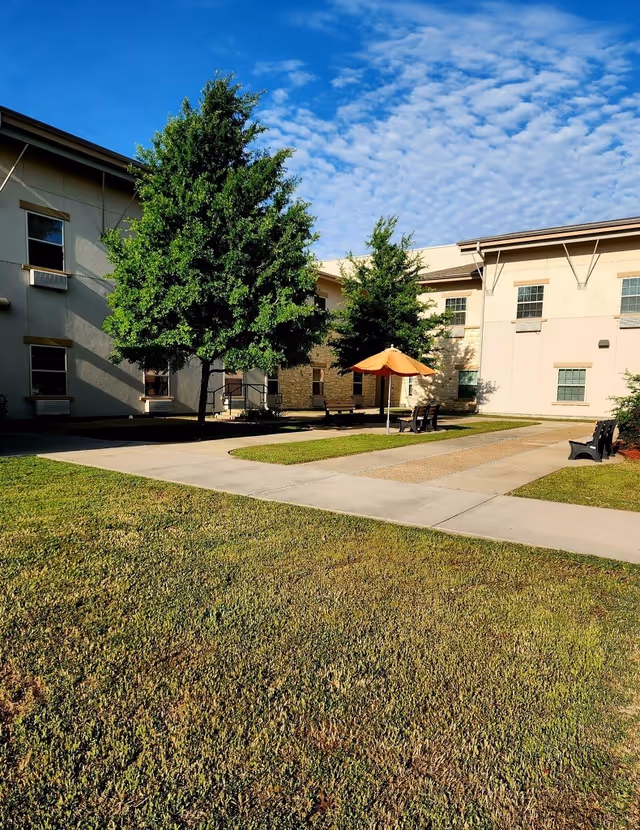 Courtyard of a senior living facility with a grassy lawn, paved walkways, trees, benches and an orange umbrella next to a two-story building under a blue sky.