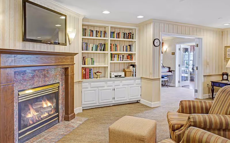 A cozy living room area featuring a lit fireplace with a wooden mantle, built-in white bookshelves filled with books and decorative items, a striped armchair, and a beige ottoman. The room has beige carpet and wallpaper with vertical stripes. A doorway leads to another room with a piano and glass doors.