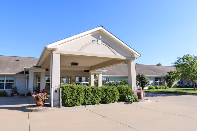 Front entrance of a senior living building with a covered porte-cochere, shrubs, and a clear blue sky.