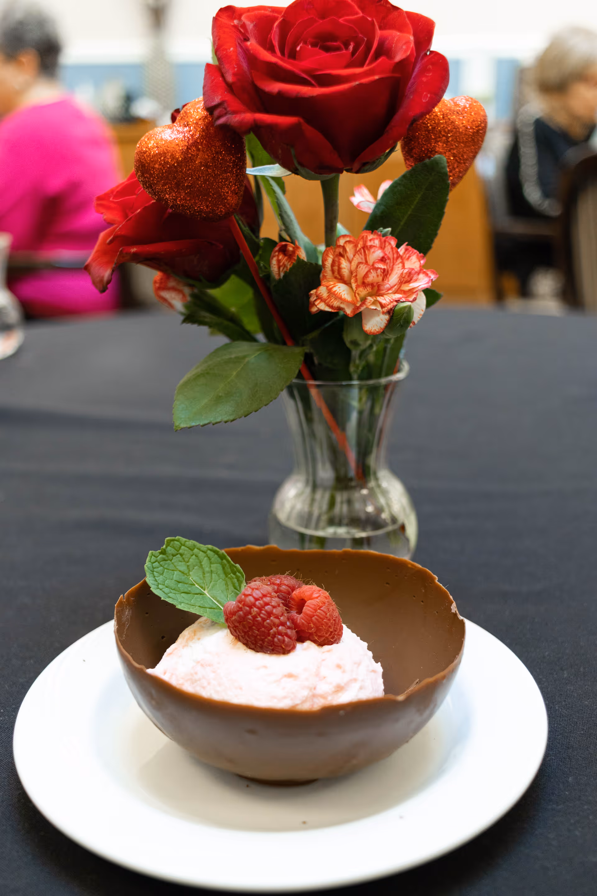 A scoop of pink ice cream topped with raspberries and a mint leaf served in a chocolate bowl on a white plate with a vase of red roses in the background.