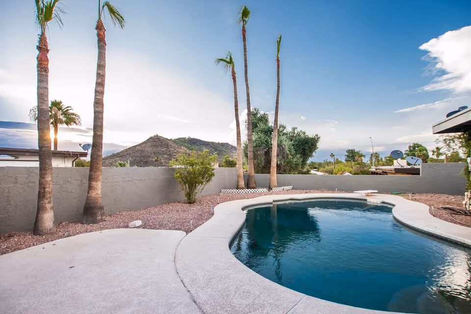 Outdoor view of a swimming pool surrounded by a concrete deck, with several tall palm trees and a low gray wall enclosing the area. In the background, there are hills and a partly cloudy sky.
