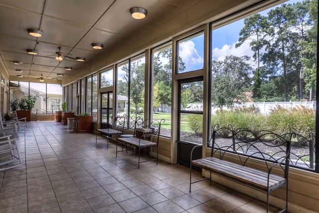 A long, tiled indoor corridor with large floor-to-ceiling windows on one side showing a view of trees and greenery outside. The corridor has several metal benches with wooden seats along the windowed wall and white rocking chairs on the opposite side. The ceiling has multiple round light fixtures.