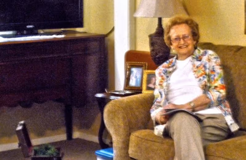 An elderly woman with glasses sitting on a brown upholstered armchair in a living room. She is smiling and holding a magazine or book. Behind her is a side table with framed photos and a lamp. A dark wooden cabinet with a television on top is visible in the background.