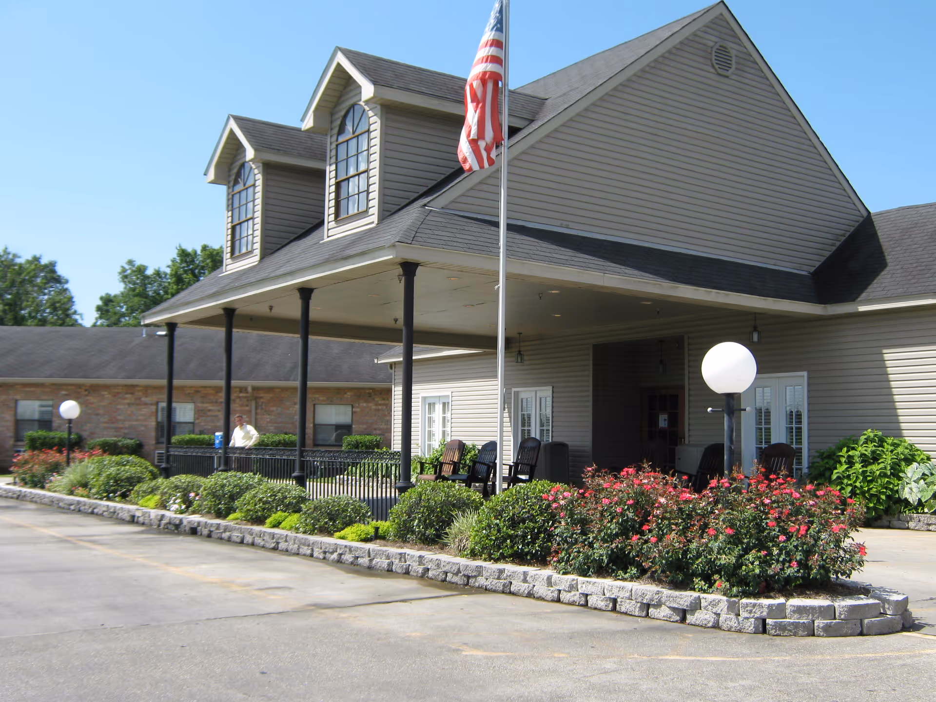Exterior view of Ormond Nursing & Care Center showing the entrance with a covered porch supported by black columns. There are two dormer windows on the roof, an American flag on a flagpole, and landscaped bushes and flowers along the driveway. Several black chairs are placed on the porch.