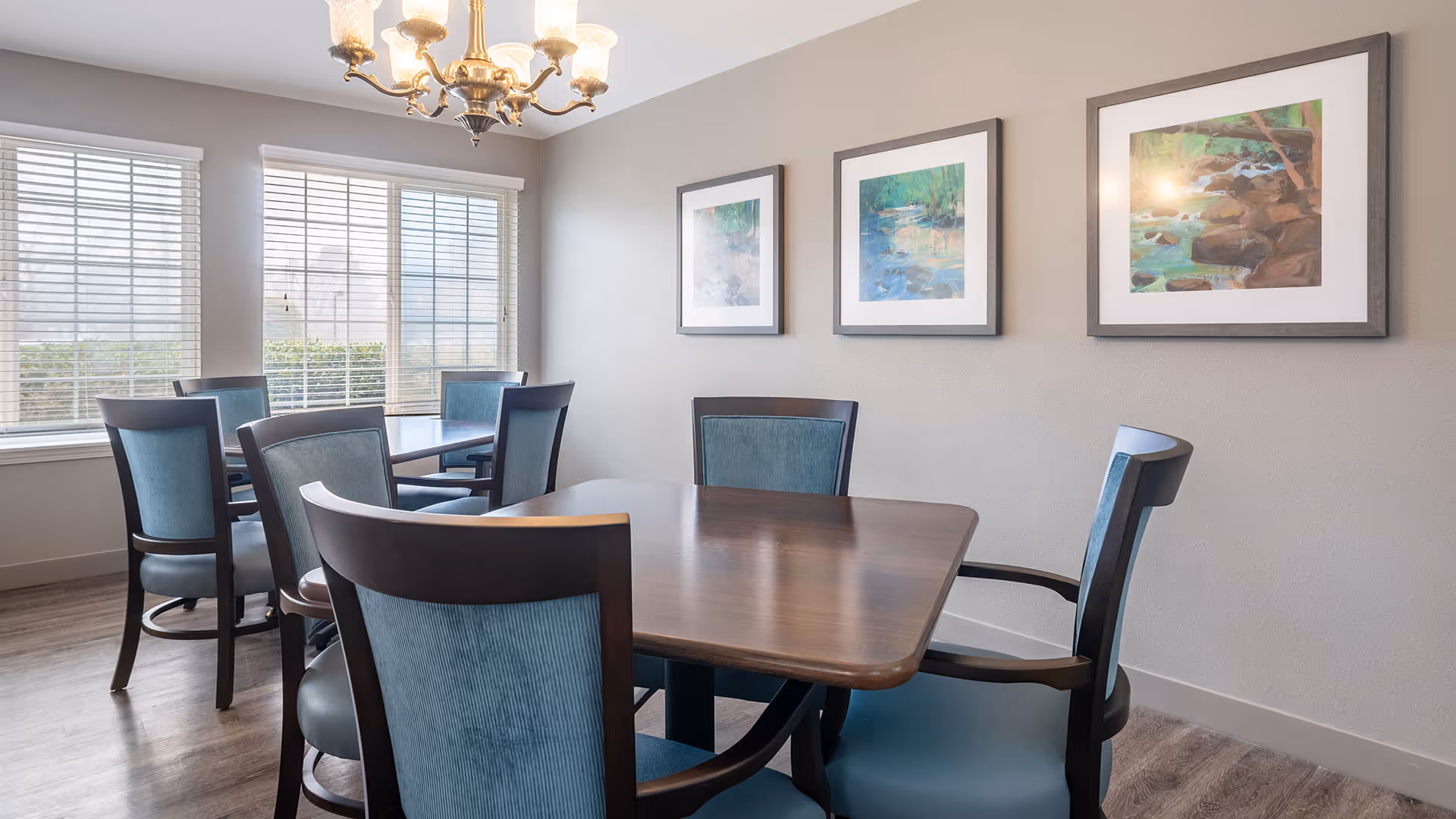 A bright dining room with wooden tables and blue cushioned chairs. Three framed paintings of nature scenes hang on a light gray wall. Large windows with white blinds allow natural light to fill the room. A brass chandelier with multiple lights hangs from the ceiling.