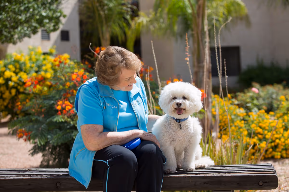 An elderly woman wearing a blue jacket and black pants sits on a wooden bench outdoors, smiling and looking at a small white fluffy dog sitting beside her. The background features colorful flowers and greenery.