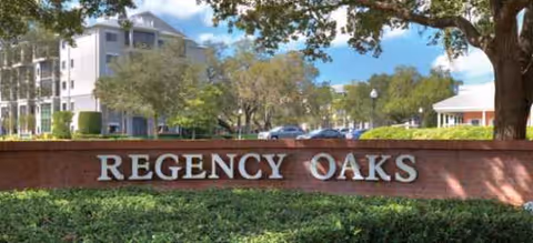 Entrance sign reading Regency Oaks with brick wall, landscaping, trees, and a building behind.