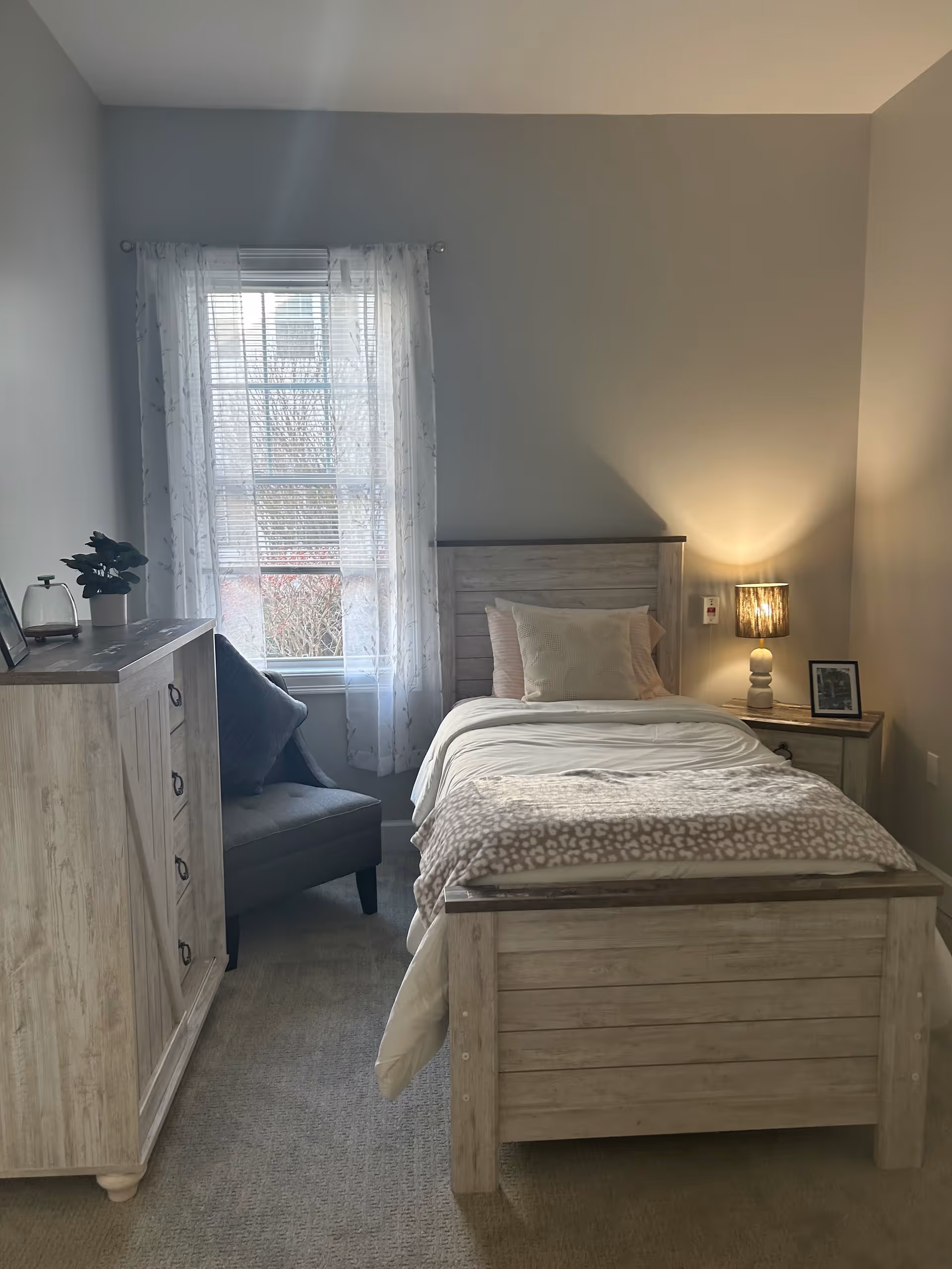 A cozy bedroom with a single wooden bed made with white and beige bedding. Next to the bed is a wooden nightstand with a lamp and a small framed picture. A wooden dresser is on the left side of the room, and a gray cushioned chair is positioned near a window with sheer white curtains, allowing natural light to enter the room.