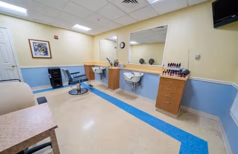 Interior of a salon room in an assisted living facility with a salon chair, two sinks with mirrors, a cabinet with nail polish bottles, a small table, and a couch. The walls are painted yellow and blue, and there is a framed picture on the wall.