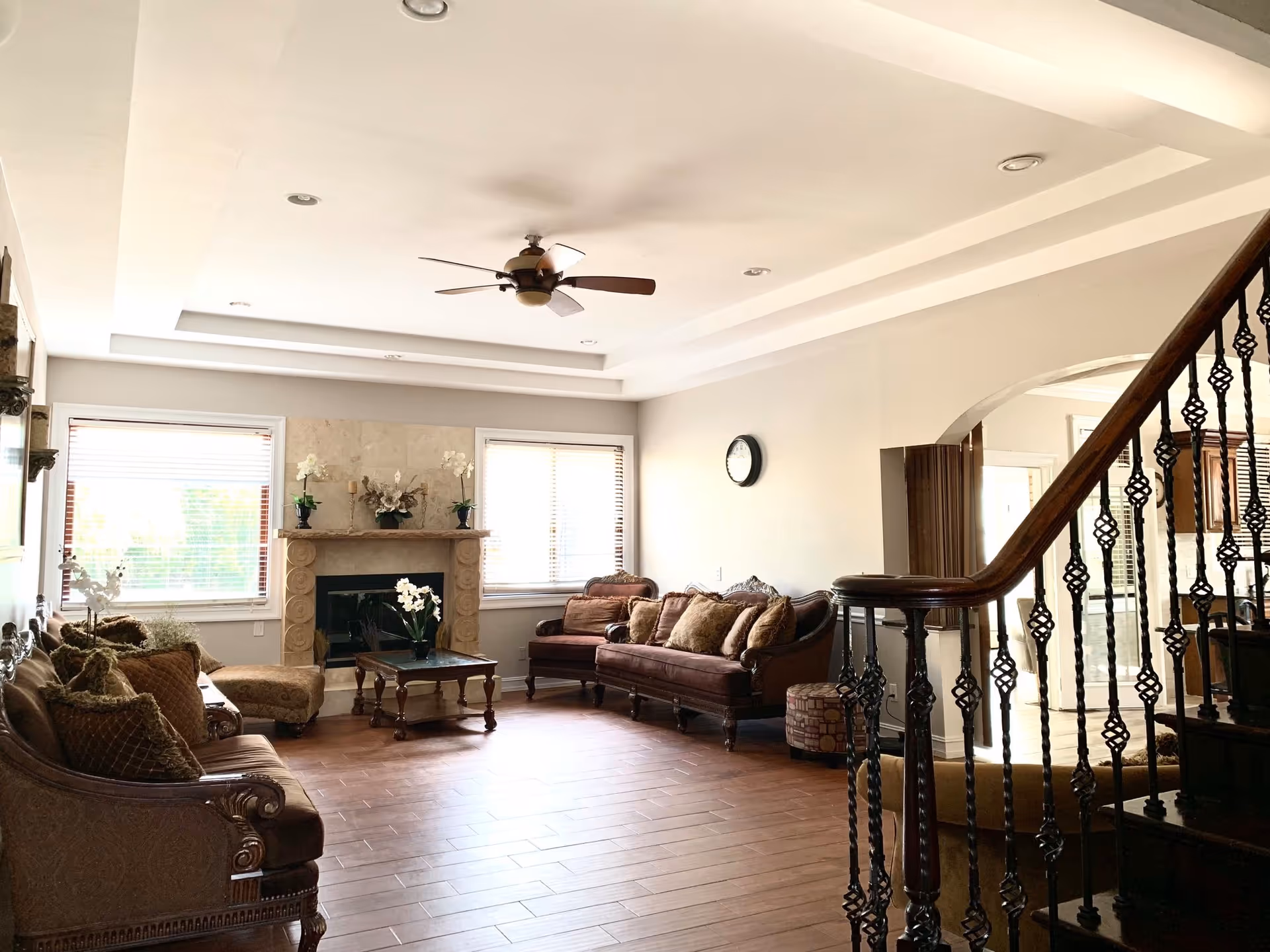 Bright living room with sofas, a fireplace, ceiling fan, large windows, and a staircase with decorative wrought-iron railing.