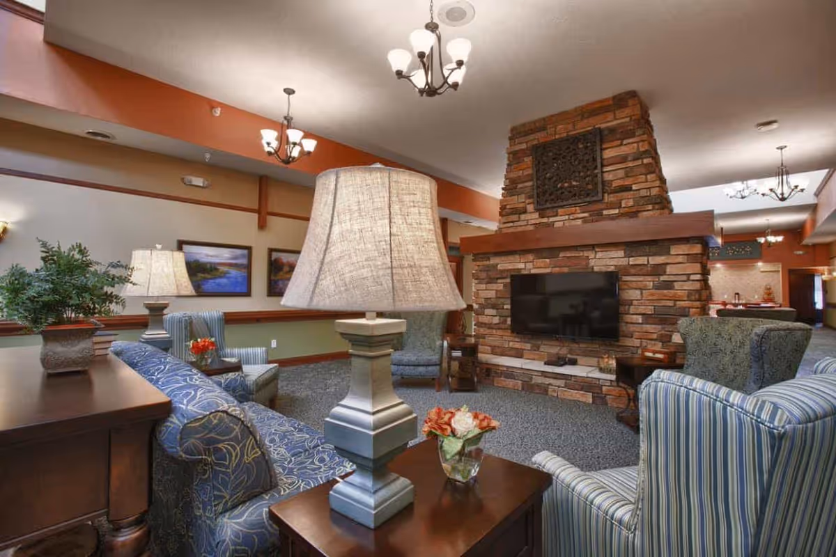 A cozy senior living facility common area with patterned armchairs and sofas arranged around a large stone fireplace with a mounted flat-screen TV. The room features warm lighting from ceiling chandeliers and table lamps, decorative plants, and framed landscape paintings on the walls.