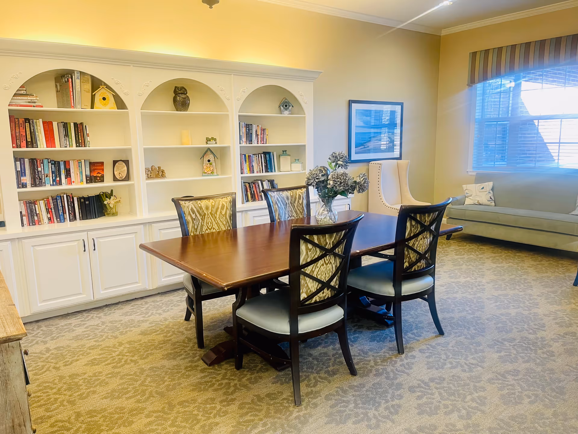 A cozy interior room with a wooden dining table surrounded by six chairs, a vase with flowers on the table, a built-in white bookshelf filled with books and decorative items, a beige armchair, a framed picture on the wall, a large window with blinds, and a light green sofa with pillows.