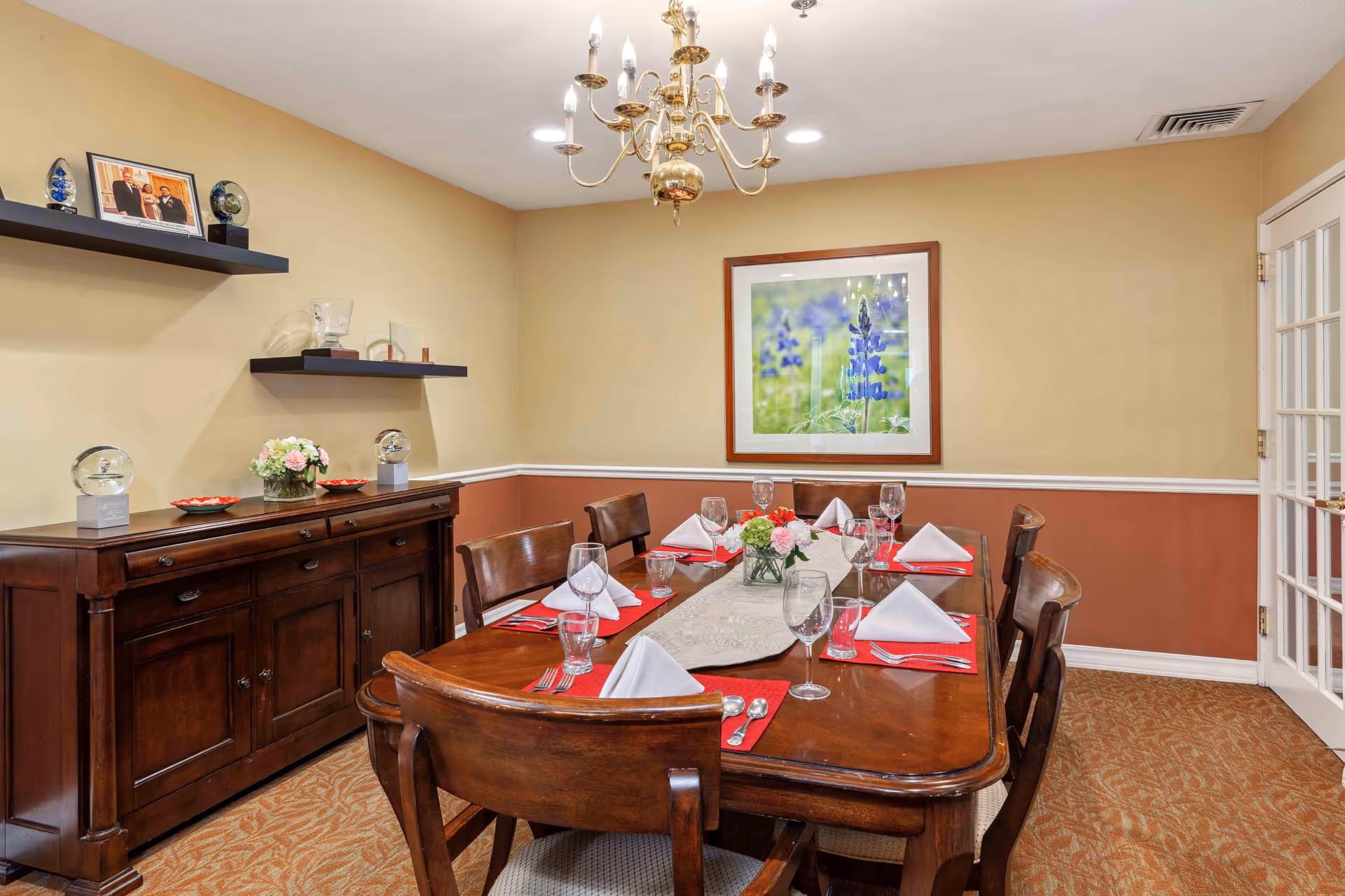 A dining room with a wooden table set for six people with white napkins, red placemats, glassware, and silverware. A floral centerpiece is in the middle of the table. The room has beige and reddish-brown walls, a chandelier hanging from the ceiling, a framed picture of purple flowers on the wall, and a wooden sideboard with decorative items and shelves above it.
