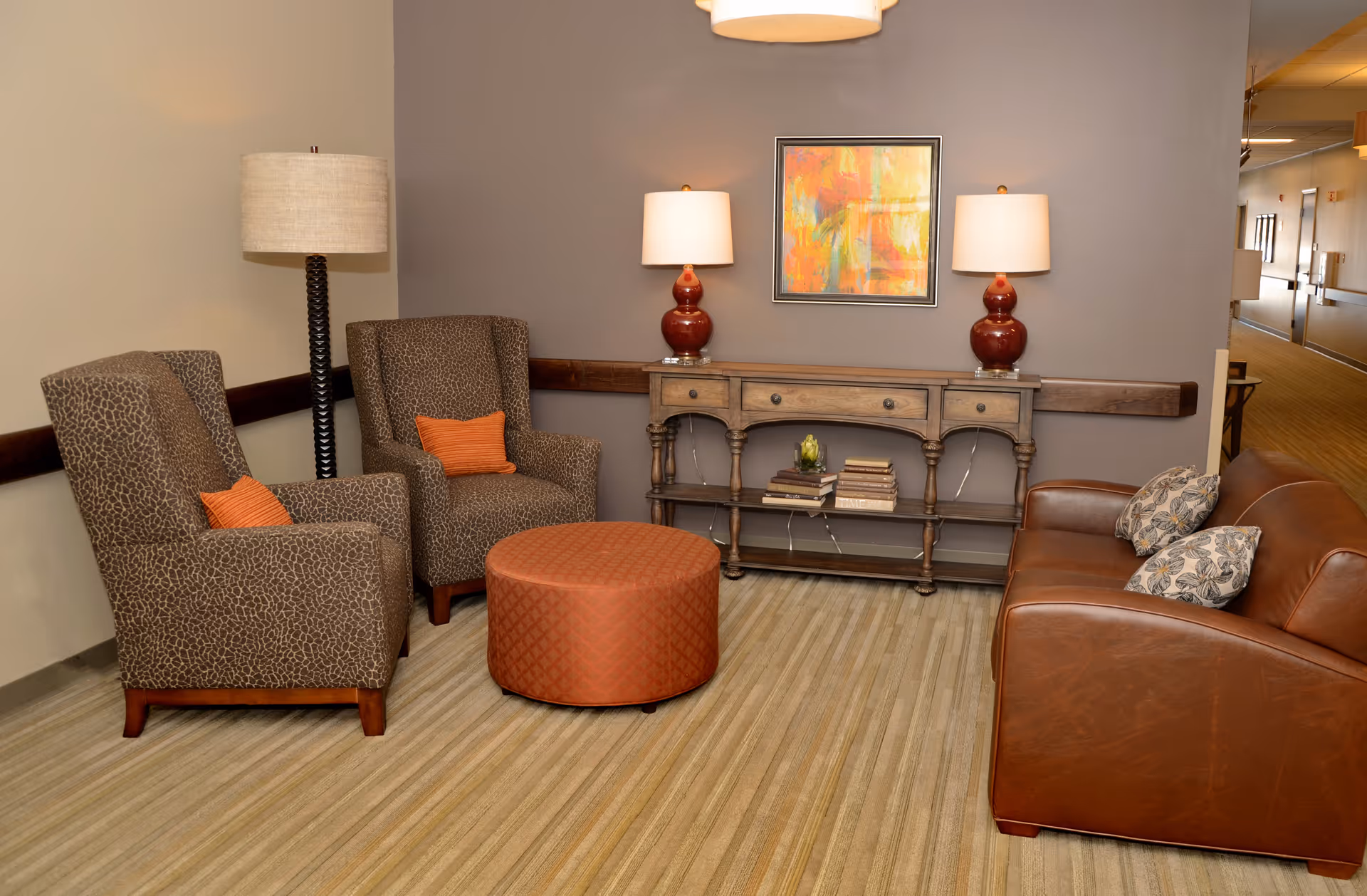 A cozy lounge seating area with two patterned armchairs, an orange ottoman, a leather sofa, and a console table with lamps and artwork.