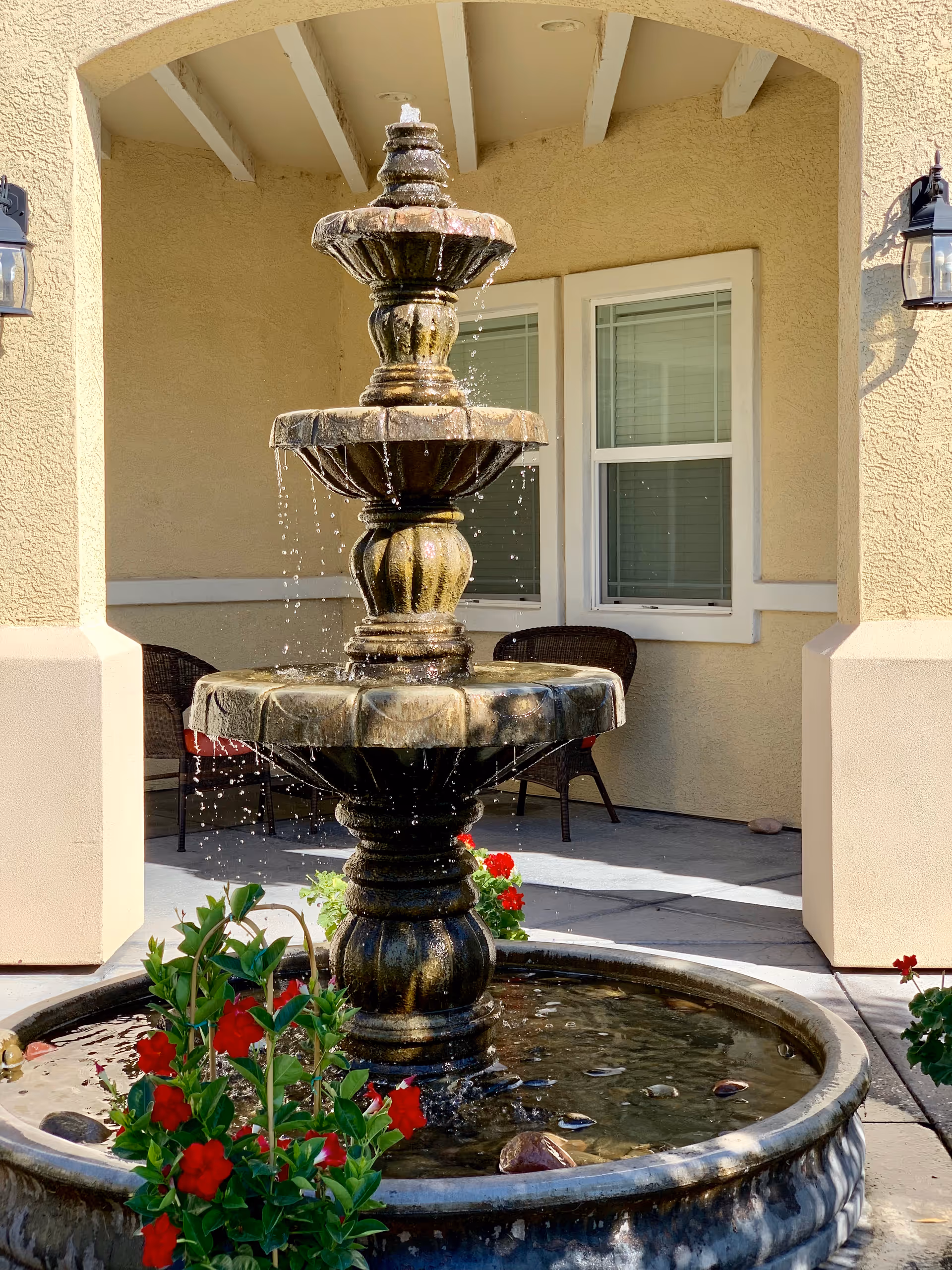 A three-tier stone fountain with water and red flowers in front of a beige building porch with windows and chairs.