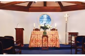 Interior view of a small chapel or worship room with a wooden lectern on the left, an altar covered with a patterned cloth and decorated with flowers and candles in the center, a tall candle stand with a cross on the right, and several chairs arranged facing the altar.