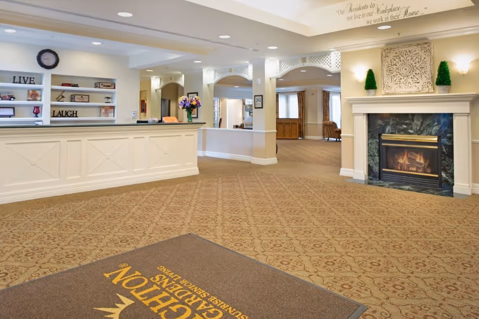Interior view of a senior living facility lobby featuring a reception desk with decorative shelves behind it, a lit fireplace with ornamental decor above, and a carpeted floor with a mat displaying the facility's name. The space is well-lit with ceiling lights and has a warm, welcoming atmosphere.