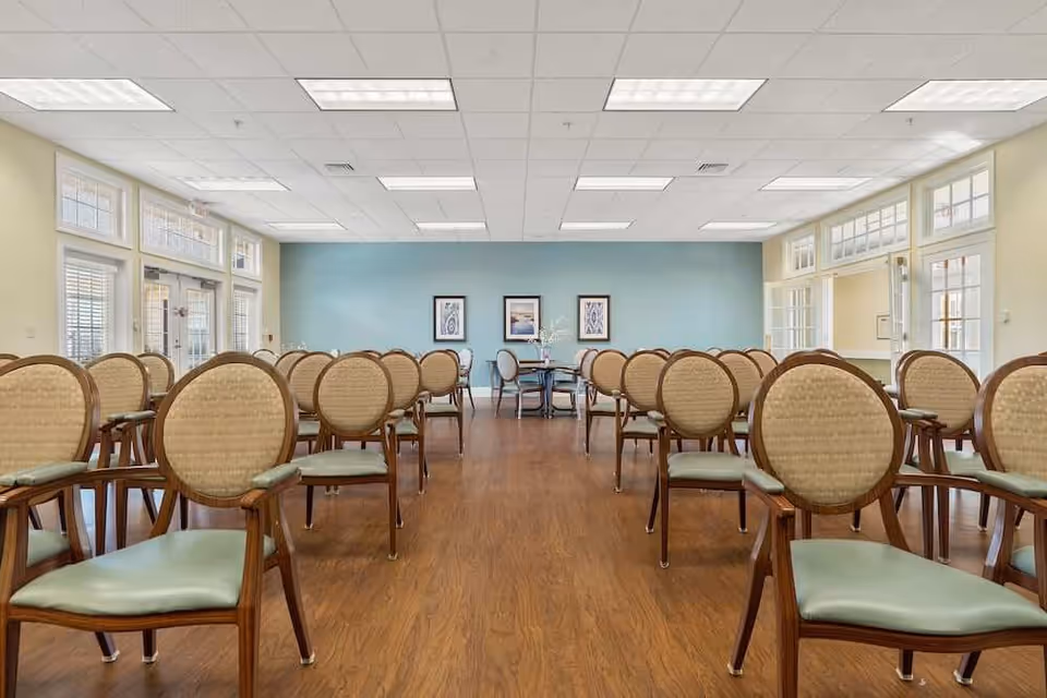 A spacious room with rows of wooden chairs featuring light green cushions and woven backs, arranged facing a blue accent wall with three framed pictures. The room has large windows and glass doors on both sides, allowing natural light to fill the space, and a wooden floor with a white ceiling featuring recessed lighting.