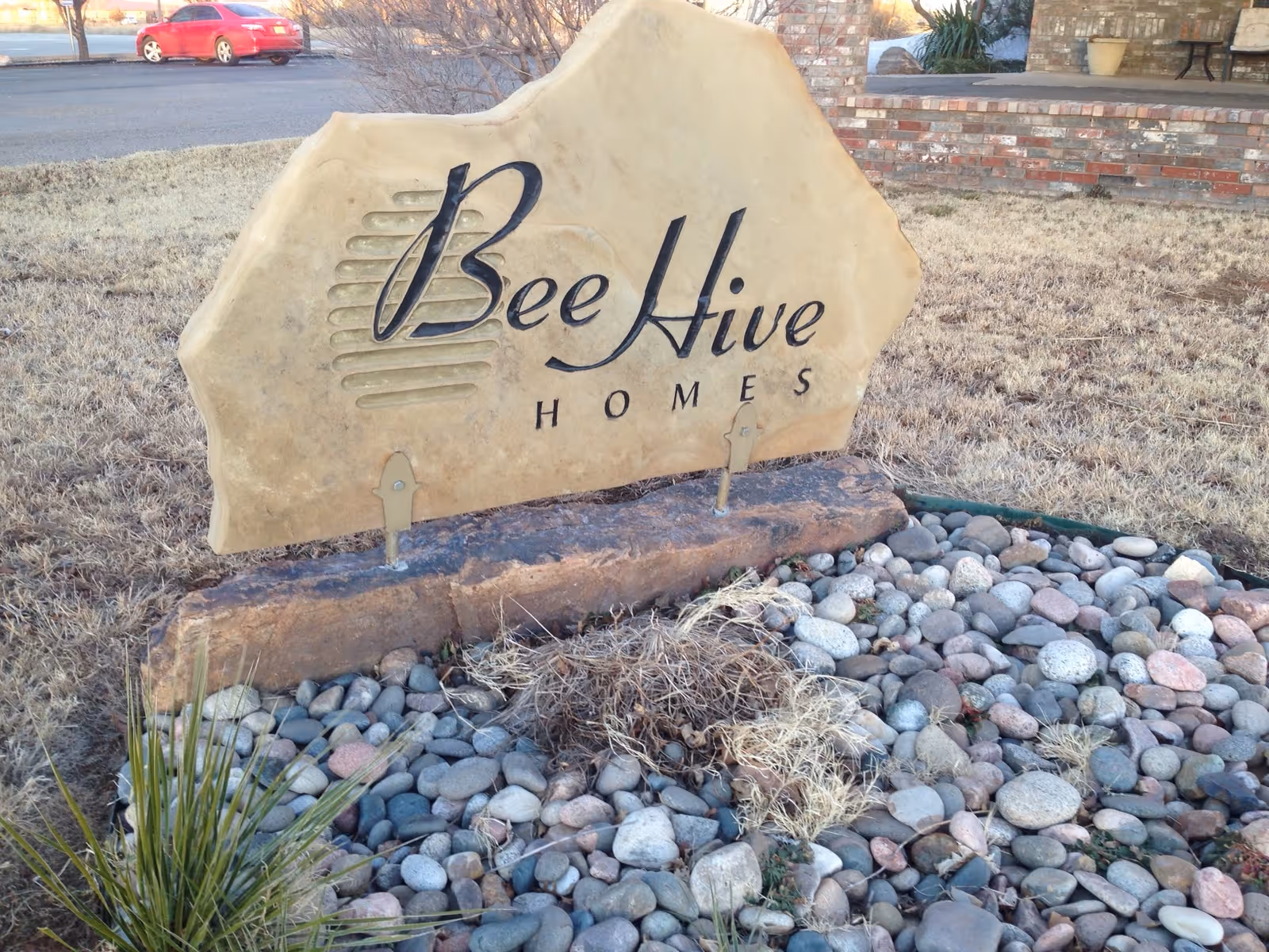Stone sign that reads 'Bee Hive Homes' on a landscaped rock bed outside a building.