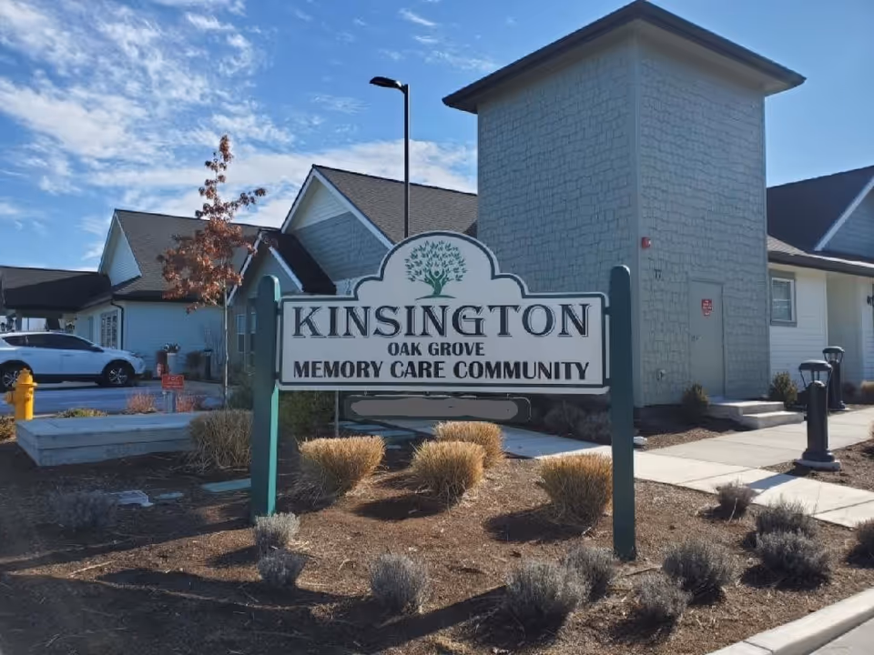 Outdoor view of the Kinsington Oak Grove Memory Care Community sign in front of a building with a clear blue sky and some landscaping including bushes and a tree.