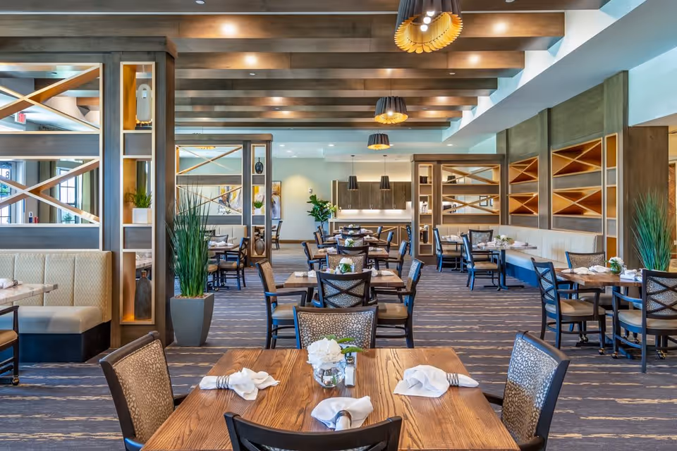 A spacious and well-lit dining room in a senior living facility with multiple wooden tables and cushioned chairs arranged neatly. The tables are set with white napkins and small floral centerpieces. The room features wooden beams on the ceiling, decorative shelving, and modern pendant lights hanging above. There are plants placed strategically around the room, and the overall decor is warm and inviting.