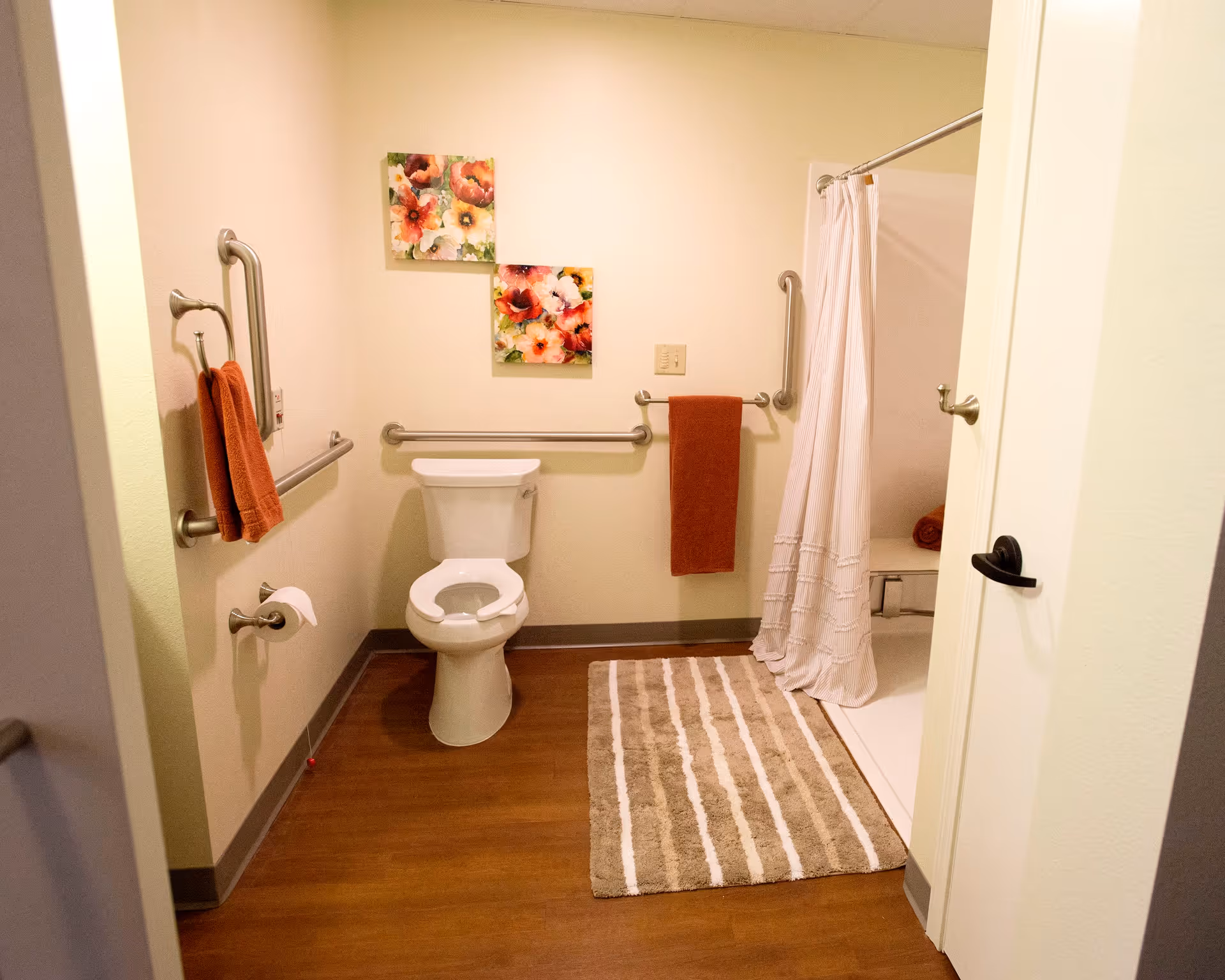 A clean and accessible bathroom with a toilet, grab bars on the walls, a towel rack with an orange towel, a striped bath mat on the wooden floor, and a shower area with a white curtain. Two floral paintings hang on the wall above the toilet.