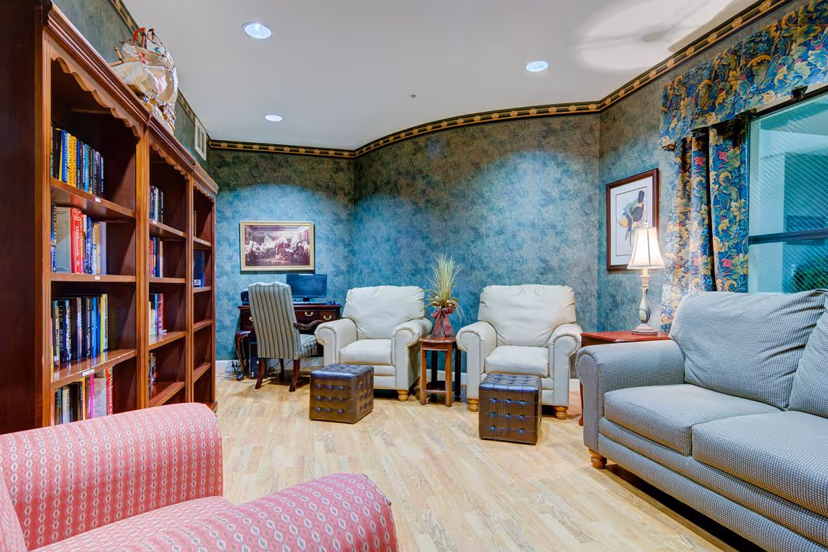 A cozy living room with wooden flooring, two white armchairs with brown ottomans, a gray sofa, a red patterned armchair, a wooden bookshelf filled with books, a desk with a computer and a striped chair, blue textured walls with framed artwork, and a window with floral curtains.