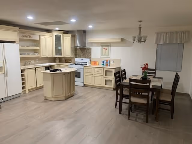 A spacious kitchen and dining area featuring cream-colored cabinets, a white refrigerator, a stove with a range hood, and a small kitchen island. The dining area has a wooden table with six chairs and a chandelier above it. The room has light wood flooring and a window with a white valance.