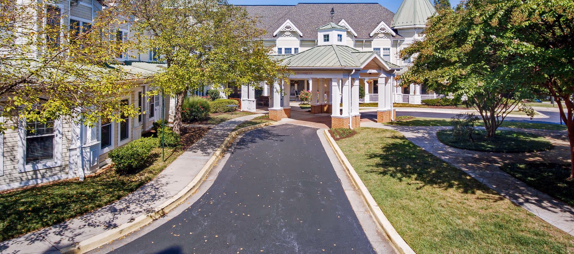 Front exterior view of Sunrise of Annapolis facility showing a covered entrance with white columns and a green roof, surrounded by trees, shrubs, and a paved driveway.