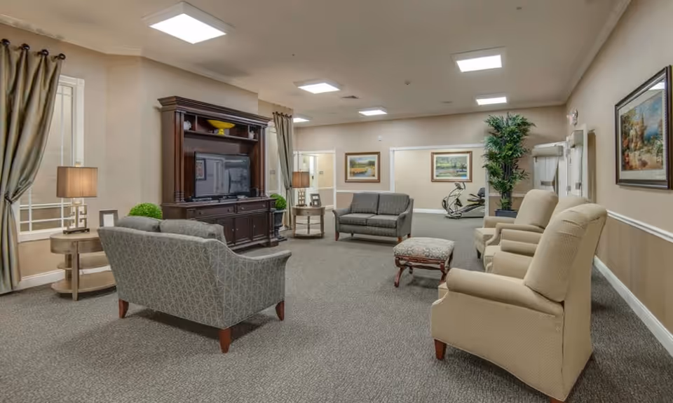 A spacious senior living common area with beige walls and carpeted floor. The room features multiple upholstered chairs and sofas arranged around a large dark wooden entertainment center with a flat-screen TV. There are two side tables with lamps, framed artwork on the walls, a large potted plant, and exercise equipment visible in the background.