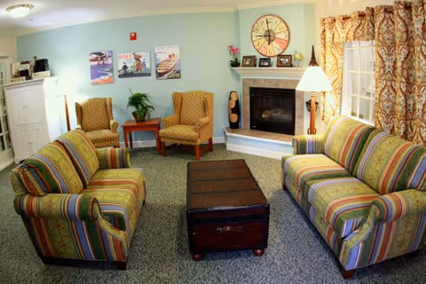 A cozy living room with two striped sofas facing each other across a dark wooden coffee table. Two patterned armchairs are placed against a light blue wall with three framed pictures above them. A fireplace with a large decorative clock and small framed photos on the mantel is on the right side. A floor lamp and a window with patterned curtains are also visible.