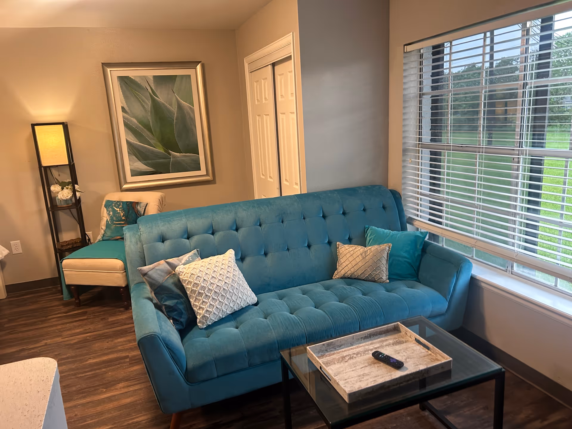 A cozy living room with a teal tufted sofa adorned with decorative pillows, a glass coffee table with a wooden tray and remote control, a beige armchair with a teal throw, a tall floor lamp, a framed close-up photo of a green succulent plant on the wall, and a large window with white blinds showing a green outdoor view.