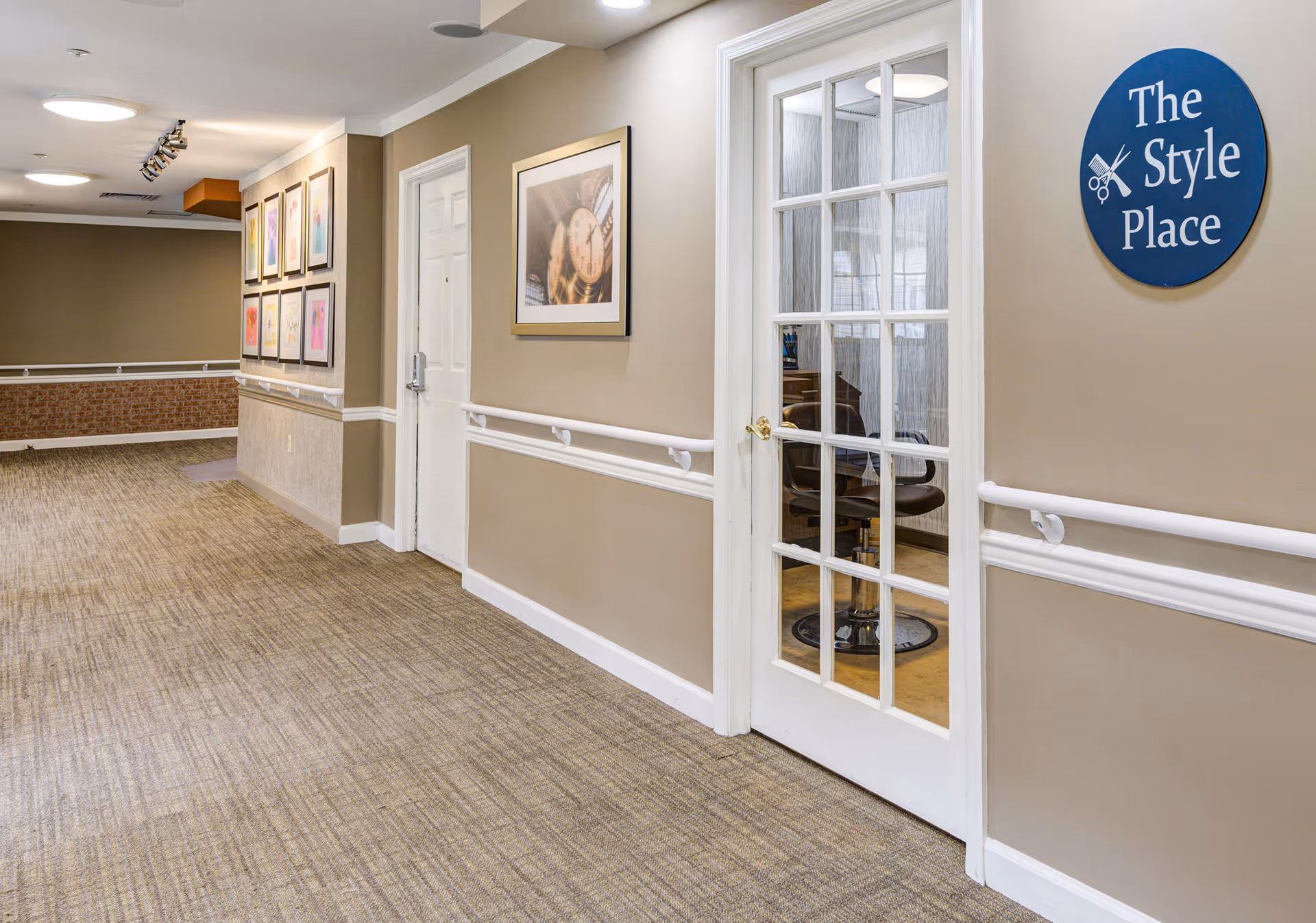Interior hallway of Arden Courts - ProMedica Memory Care Community with beige walls, carpeted floor, white handrails, framed artwork on the walls, and a glass door leading to a room labeled 'The Style Place' with a barber chair visible inside.