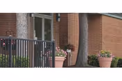 Brick building entrance with glass double doors, potted plants, a black metal railing, and nearby trees.