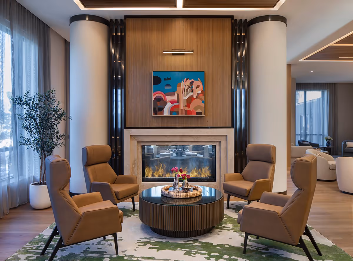 Cozy lounge with four brown high-backed chairs around a round coffee table facing a modern fireplace topped by colorful abstract artwork.