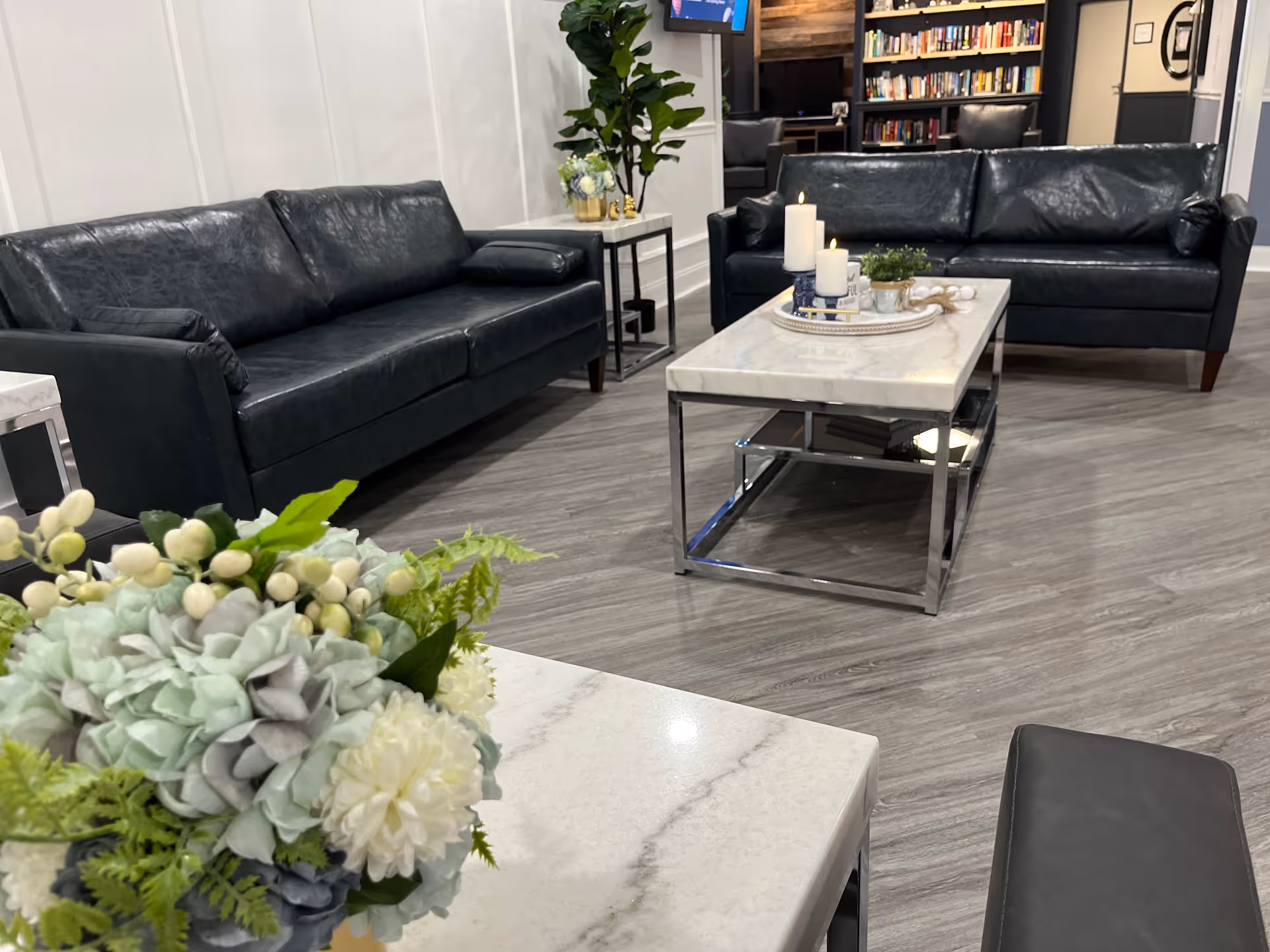A modern living room area with two black leather sofas facing a rectangular marble coffee table. The coffee table has decorative candles and a small plant on it. In the foreground, there is a white marble side table with a floral arrangement. The room has wood flooring, white paneled walls, and a bookshelf with books and a TV mounted above it in the background.