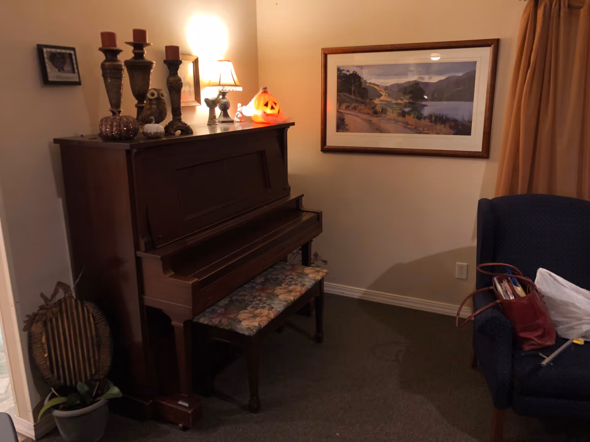 Upright piano with a floral bench and decorative items on top, a framed landscape on the wall, and an armchair with a bag.