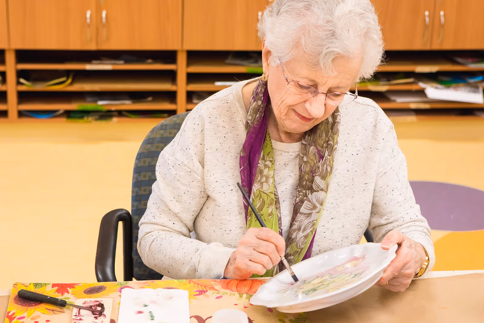 An elderly woman with white hair and glasses is sitting at a table painting a decorative plate. She is wearing a light-colored sweater and a floral scarf. The background shows wooden shelves with various papers and materials.