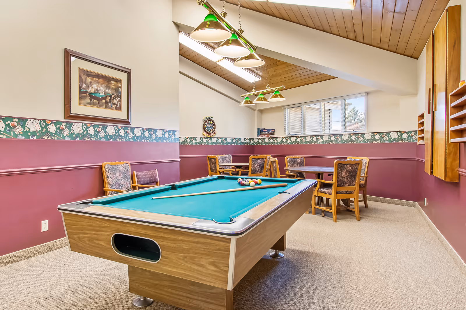 A recreational room with a pool table in the center, a cue stick and billiard balls arranged on the table. The room has maroon walls with a decorative wallpaper border featuring playing cards and dice. There are several wooden chairs with floral upholstery and round tables along the walls. The ceiling is wooden with skylights and hanging green-shaded lights above the pool table. A framed picture and a round wall clock with the words 'Pool Billiards' are visible on the walls.