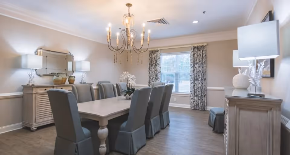 A well-lit dining room with a long rectangular table surrounded by eight upholstered chairs. The room features a chandelier hanging above the table, two sideboards with decorative lamps and vases, a large window with patterned curtains, and light-colored walls with crown molding.