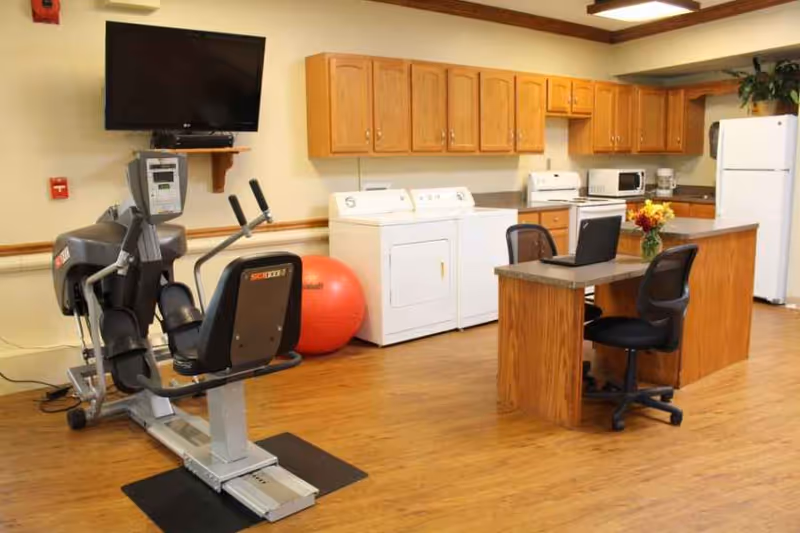 A room with exercise equipment including a recumbent bike and a large red exercise ball, adjacent to a laundry area with a washer and dryer. The room also features a kitchen area with wooden cabinets, a refrigerator, microwave, coffee maker, and a small island with two office chairs and a laptop on it.