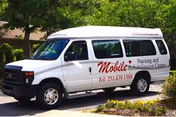 White passenger van parked on a driveway with trees and bushes in the background. The van has text on the side that reads 'Mobile Nursing and Rehabilitation Center' along with a phone number.