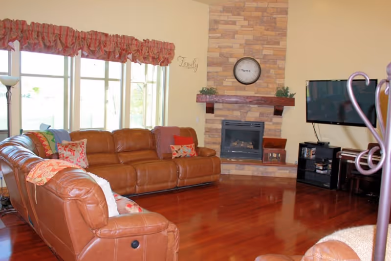 A spacious living room with a large brown leather sectional sofa adorned with colorful pillows and blankets. The room features a stone fireplace with a wooden mantle, a wall clock above it, and a flat-screen TV mounted on the wall. Large windows with red valances allow natural light to fill the room, and the floor is polished wood.