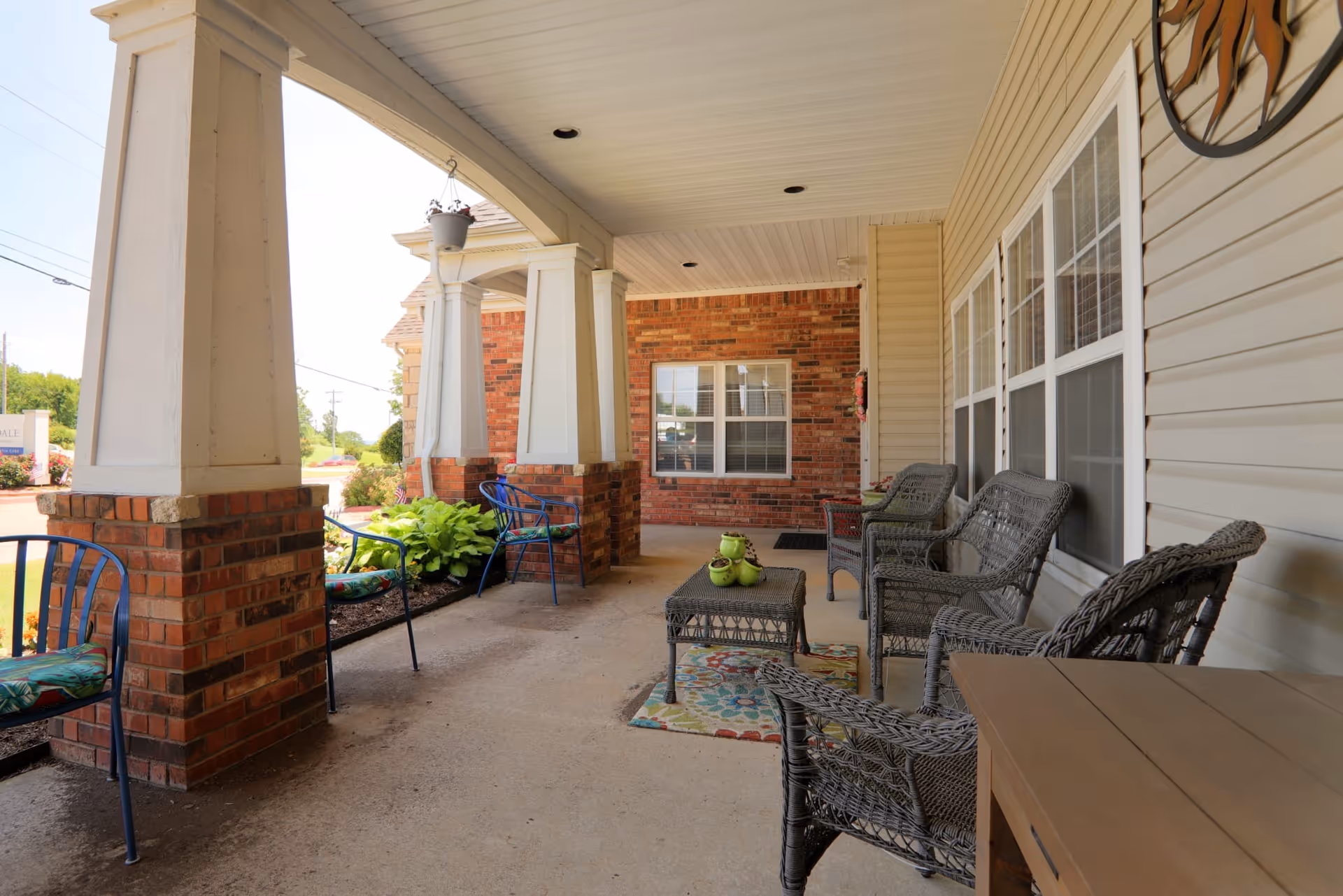 Covered outdoor porch area with brick pillars and white siding walls. The porch is furnished with wicker chairs, a small wicker table with green decorative items, metal chairs with floral cushions, and a colorful rug. There are plants and greenery along the edge of the porch and a window on the brick wall in the background.