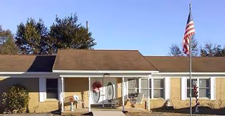 Single-story brick senior living facility entrance with a covered porch, chairs, and an American flag.